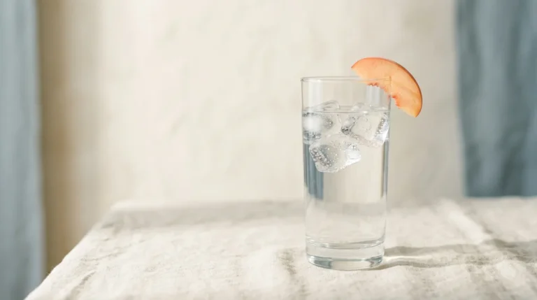 A clear glass of ice water with a thin peach slice on the rim, on a cream linen surface in soft window light